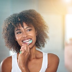 Woman in white tank top with curly brown hair brushing her teeth at a mirror