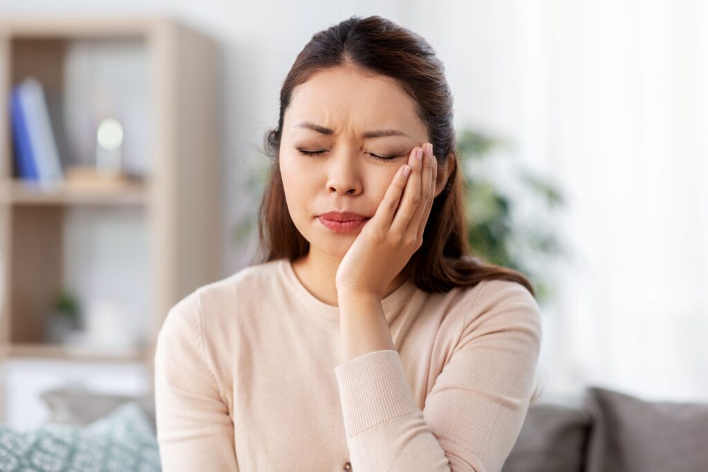 Woman in neutral sweater holding hand to jaw in pain with eyes closed